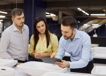 Customers consult a salesman in a plumbing showroom. A tablet helps compare faucets and sinks, with consultation on pricing for shopping selection. Guided plumbing product selection idea.