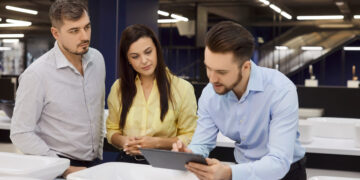 Customers consult a salesman in a plumbing showroom. A tablet helps compare faucets and sinks, with consultation on pricing for shopping selection. Guided plumbing product selection idea.